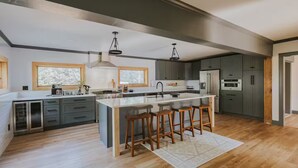 A modern kitchen with a central island, bar stools, and a large window. - Lyon Mountain Lodge (Saranac)