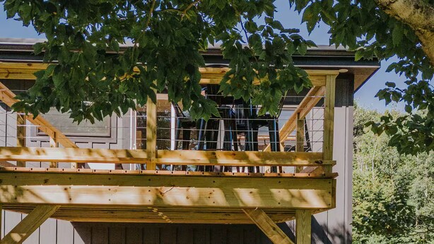 A wooden deck with outdoor seating, a glass door leading to a covered area, and a tree overhead. - Lyon Mountain Lodge (Saranac)