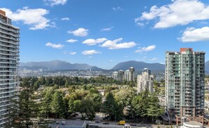 A high-rise building with multiple balconies, surrounded by trees and other buildings, with mountains in the background. - Modern 2 Bed 2 Bath Condo with Parking, Patio & Amenities – Prime Location (Surrey)