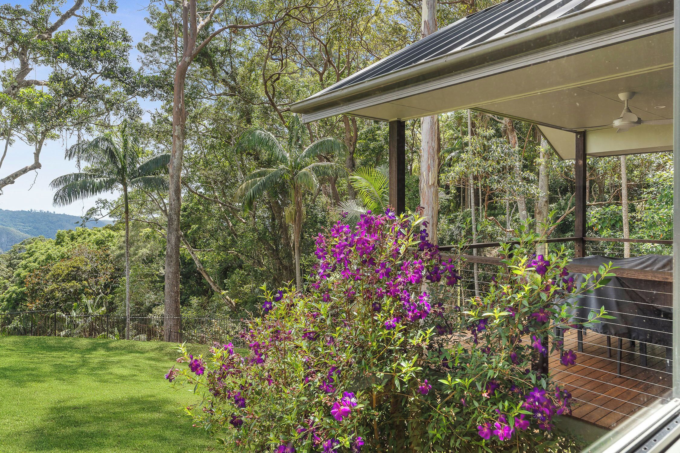 Una casa con porche cubierto, rodeada de exuberante follaje y flores moradas.