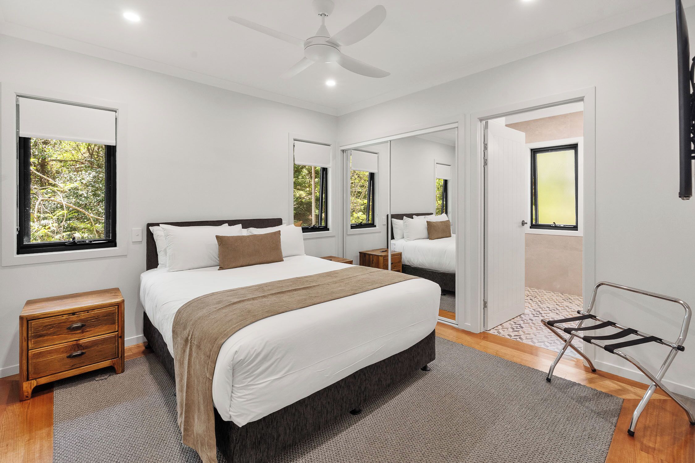 A modern bedroom with a large bed, bedside tables, a ceiling fan, and a view of the outdoors through windows.