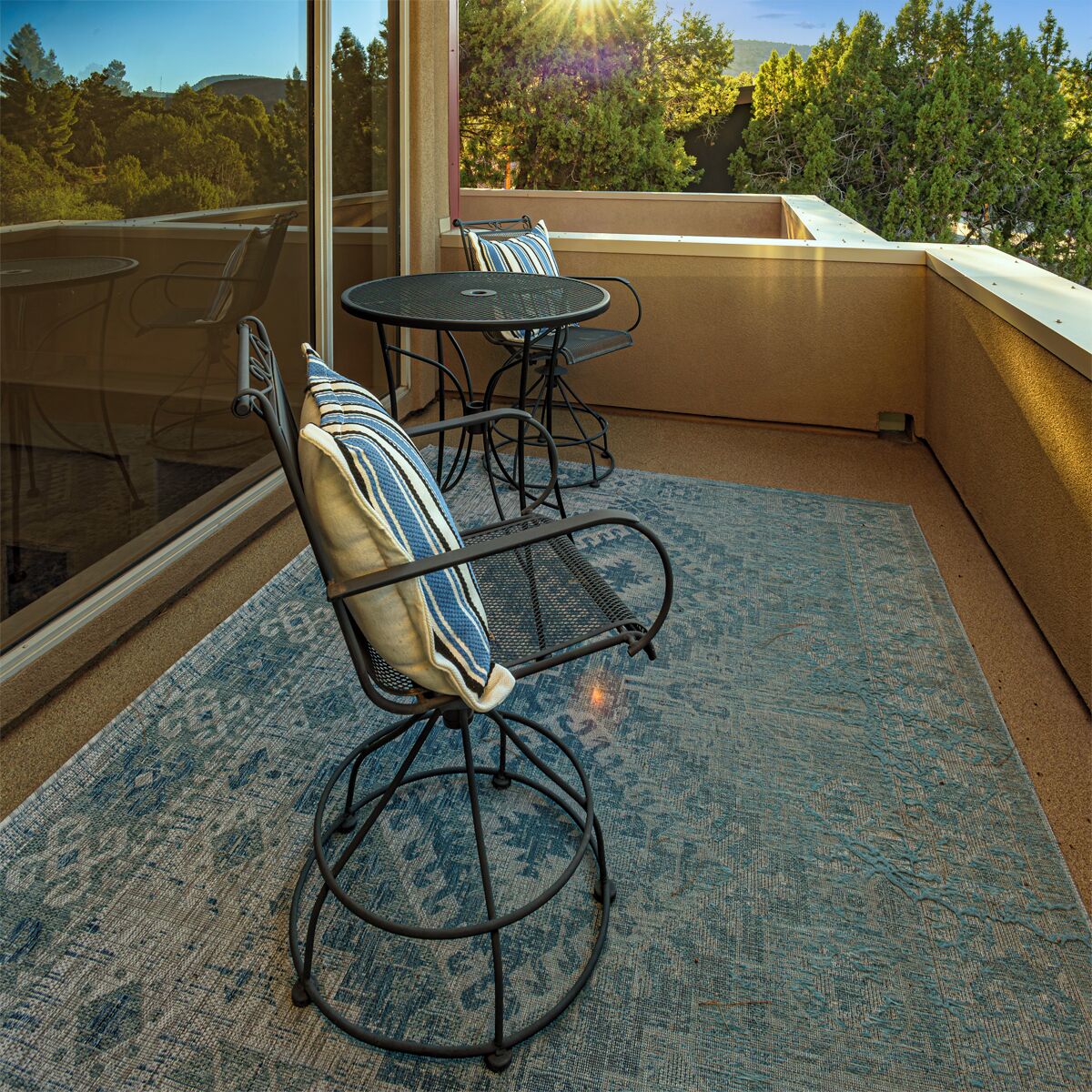 A balcony with two metal chairs and a small table, a patterned rug, and a view of trees and hills.