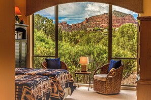 A room with a large window offering a view of red rock formations and greenery. There is a bed with patterned bedding and two wicker chairs with cushions. - Bell Rock Haven 3bd 3.5ba Hot Tub + Amazing Views (Sedona)
