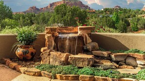 Un jardín rocoso con una pequeña cascada, rodeado de vegetación y un telón de fondo montañoso.