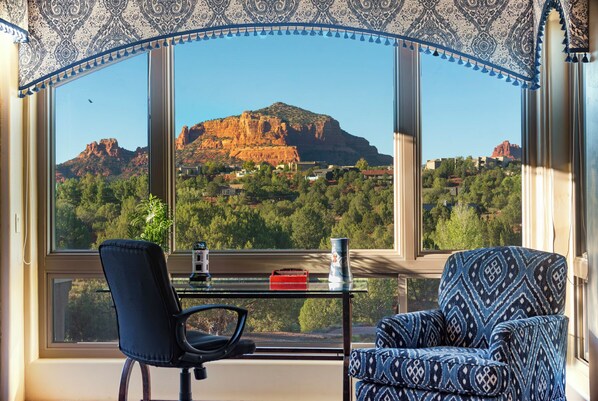 Ein Zimmer mit großem Fenster und Blick auf Berg und Wald. Es gibt einen schwarzen Bürostuhl, ein gemustertes Sofa und einen kleinen Tisch mit Vase und Uhr.