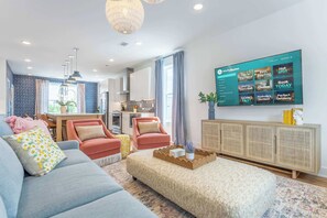 A modern living room with a flat-screen TV displaying a streaming service interface, a sofa, a coffee table, and a dining area in the background.