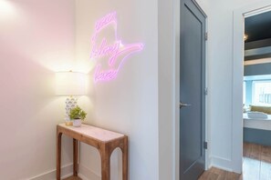 Een gang met een neonbord, een lamp, een kleine tafel met een plant en een deur naar een kamer.