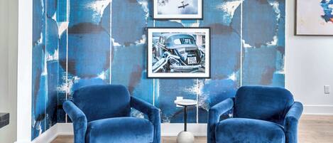 Una habitación con dos sillones tapizados en terciopelo azul, una mesita redonda pequeña y una pared decorada con motivos abstractos en azul y blanco.
