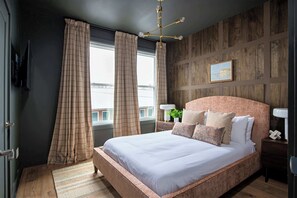 Un dormitorio con una cama grande, cabecera de madera, mesita de noche con lámpara y ventana con cortinas.
