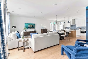 A modern living room with a sofa, a coffee table, and a blue armchair. There is a kitchen area with white cabinets and a dining table.