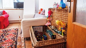 Un salon chaleureux avec un panier en osier rempli de jouets et de livres, un fauteuil rouge, un canapé blanc et un téléviseur posé sur un meuble.