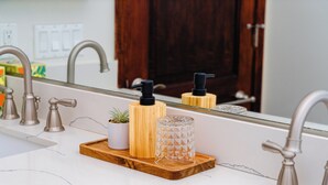 Ein Badezimmer mit einem weißen Marmorwaschbecken, einer Holztablett mit einem Blumentopf und zwei schwarzen Seifenspendern sowie einem Glaswindlicht.