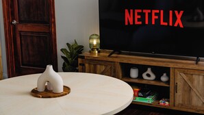 Ein Wohnzimmer mit einem TV-Schrank aus Holz, einem Flachbildfernseher, der "NETFLIX" anzeigt, einem runden Tisch mit weißer Vase und einer kleinen Pflanze.