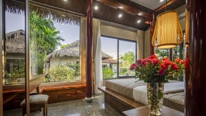 A room with a large window, a bed, a chair, a table with a vase of flowers, and a hanging lamp. - Lá Retreat (Da Nang)