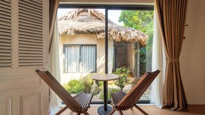 A room with two wooden chairs facing a glass door leading to a balcony with a thatched roof and a view of greenery. - Lá Retreat (Da Nang)