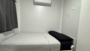 A small, white, single-bed room with a grey bedspread, a white bedside table, a grey rug, and a white air conditioning unit on the wall.
