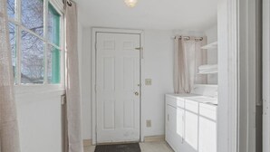 Um corredor estreito com uma porta branca, uma janela com detalhes verdes e uma área de serviço com máquina de lavar e secadora.