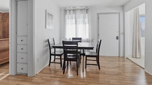 Uma área de refeições com mesa e cadeiras de madeira, paredes brancas e uma porta que dá para outro espaço.