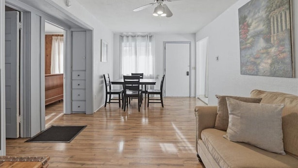 Um quarto espaçoso com piso de madeira, área de refeições com mesa e cadeiras, um ventilador de teto e um quadro na parede.