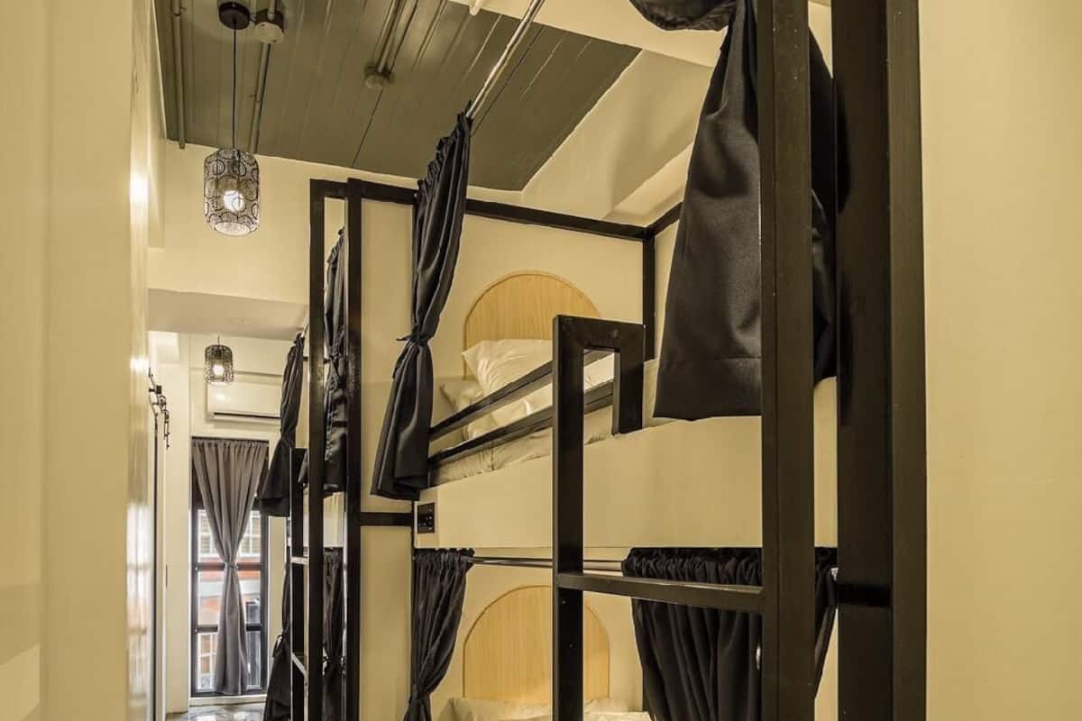 Un corridoio stretto con letti a castello su entrambi i lati, un pavimento decorato e un soffitto con tubature e luci a vista.