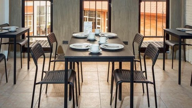 Un comedor con una mesa central preparada para seis personas, rodeada de sillas, y amplias ventanas que ofrecen vistas a un edificio y un paisaje.