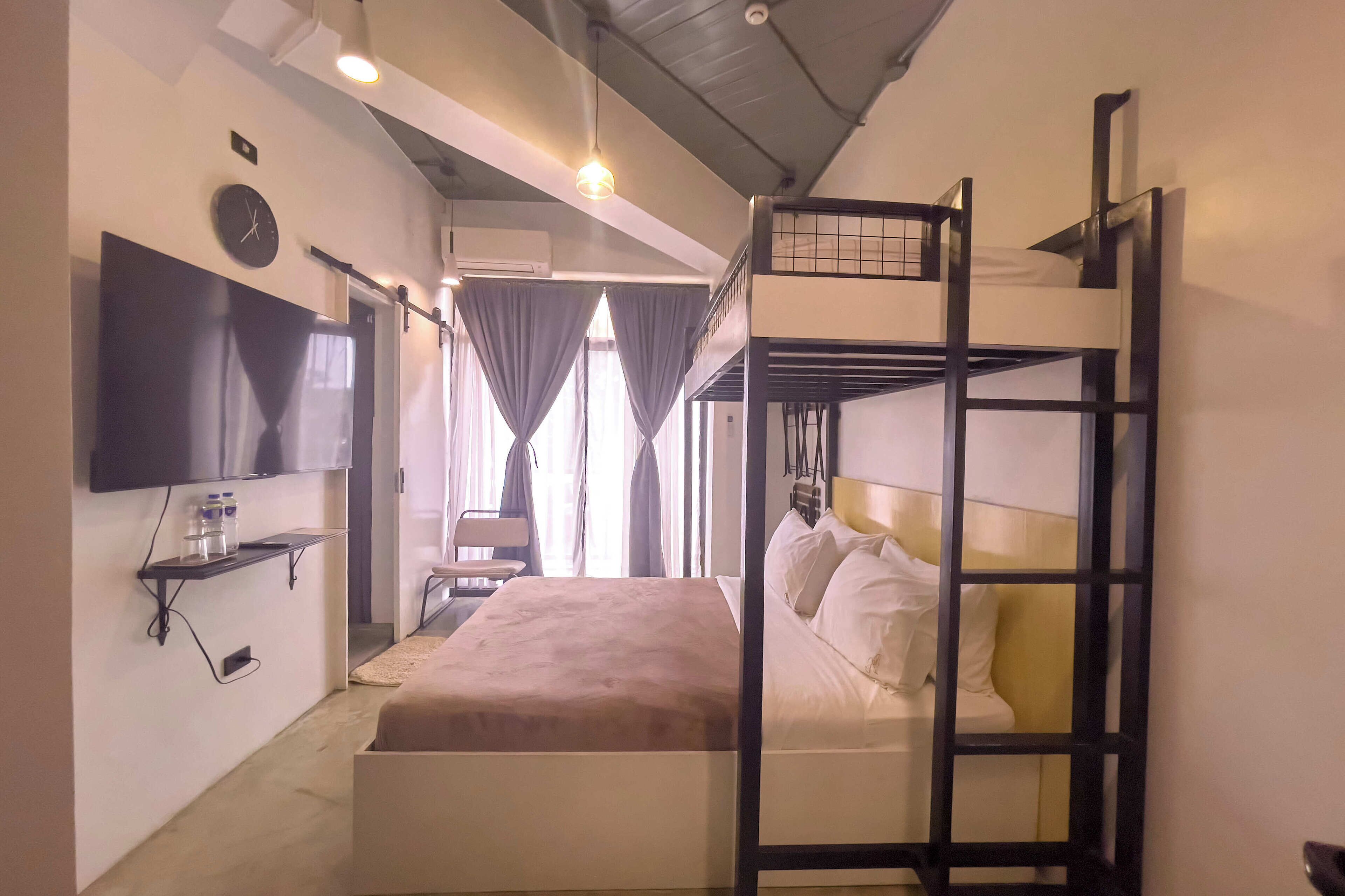 Quarto com beliche, televisão de tela plana na parede, mesa pequena com garrafa e copos, e janela com cortinas.