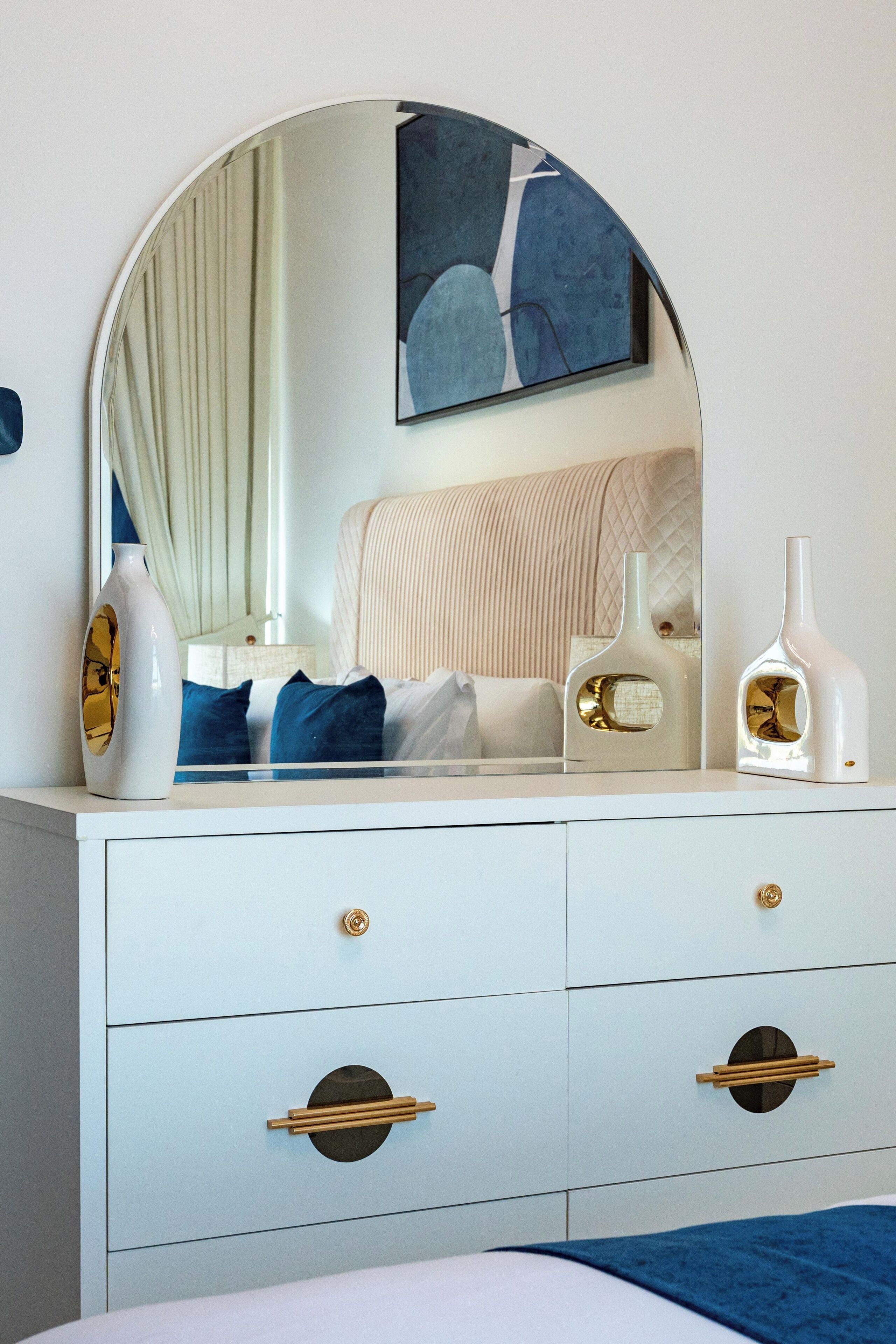 A white dresser with gold handles, a mirror, and two decorative vases.