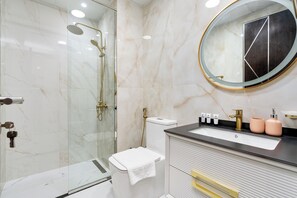 Casa de banho moderna com chuveiro envolto em vidro, espelho redondo e toucador com artigos de higiene pessoal.
