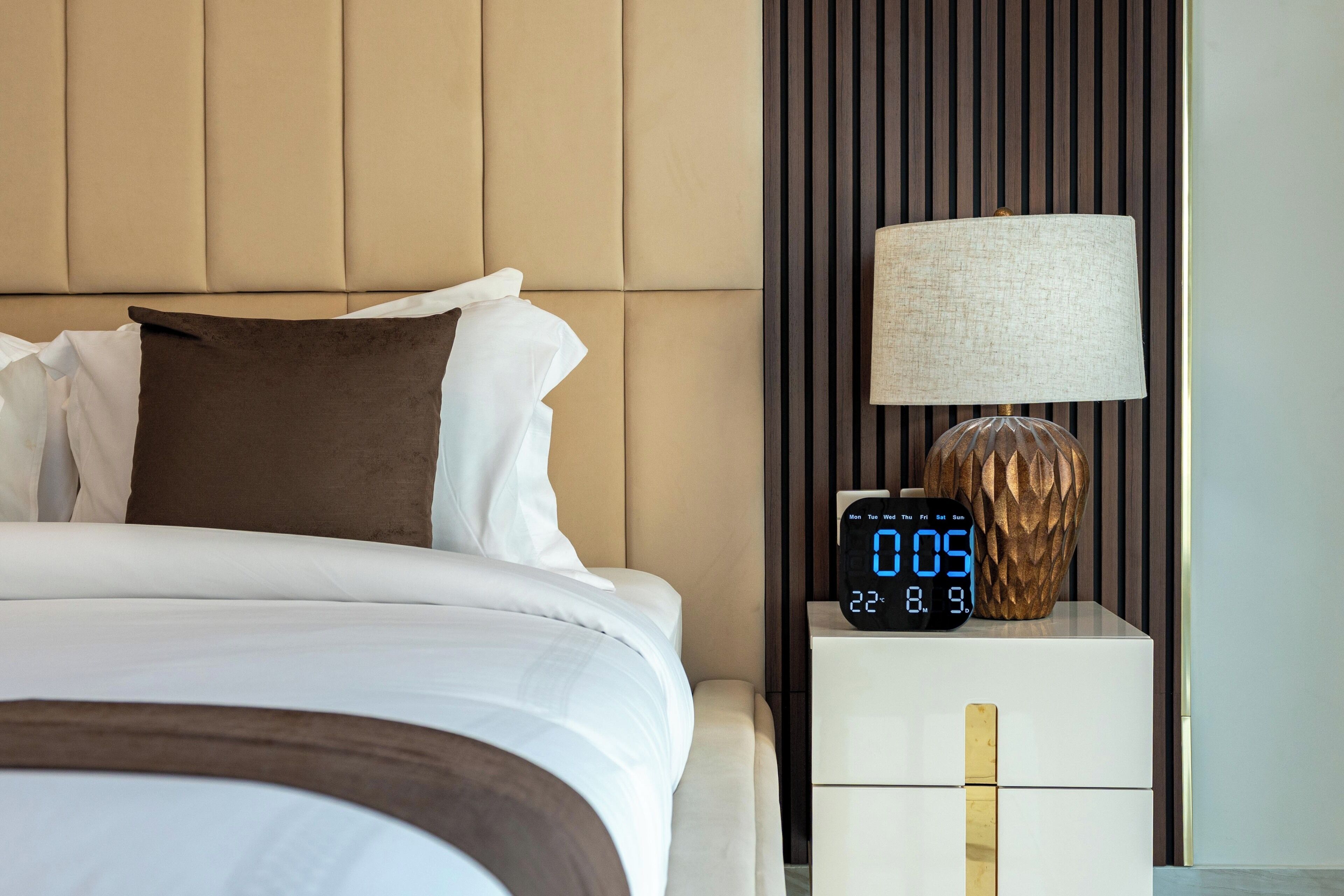 Een net opgemaakt bed met wit en bruin beddengoed, een digitale klok die de tijd toont en een lamp op een nachtkastje.