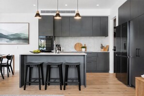 Une cuisine moderne dotée d’un îlot central, de tabourets de bar noirs, d’un coin repas avec une table et des chaises, et d’un grand réfrigérateur.