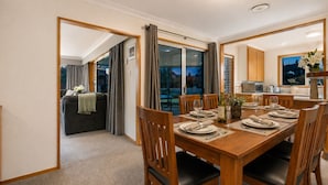 A dining area with a wooden table set for four, a kitchen in the background, and a balcony with curtains.
