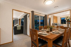 A dining area with a wooden table set for four, a kitchen in the background, and a balcony with curtains.