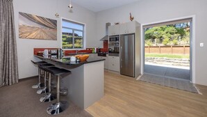 A modern kitchen with a central island, bar stools, stainless steel appliances, and a sliding glass door leading to a backyard. - Amazing views of Lake and Mountains on Bovett (Wānaka)
