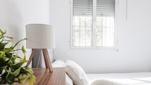 Un dormitorio minimalista con una mesita de noche de madera, una lámpara blanca, una cama con ropa de cama blanca y una ventana con persianas.
