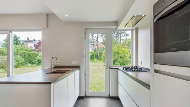 A modern kitchen with a large island, built-in oven, and a view of the garden through the door. - Renovated villa with large garden (Koksijde)