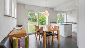 A modern kitchen with a large island, a dining area with a wooden table and chairs, and a view of a garden through the sliding glass doors. - Renovated villa with large garden (Koksijde)