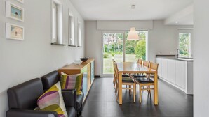 A modern kitchen with a dining area, a black leather sofa, and a large window offering a view of the garden. - Renovated villa with large garden (Koksijde)
