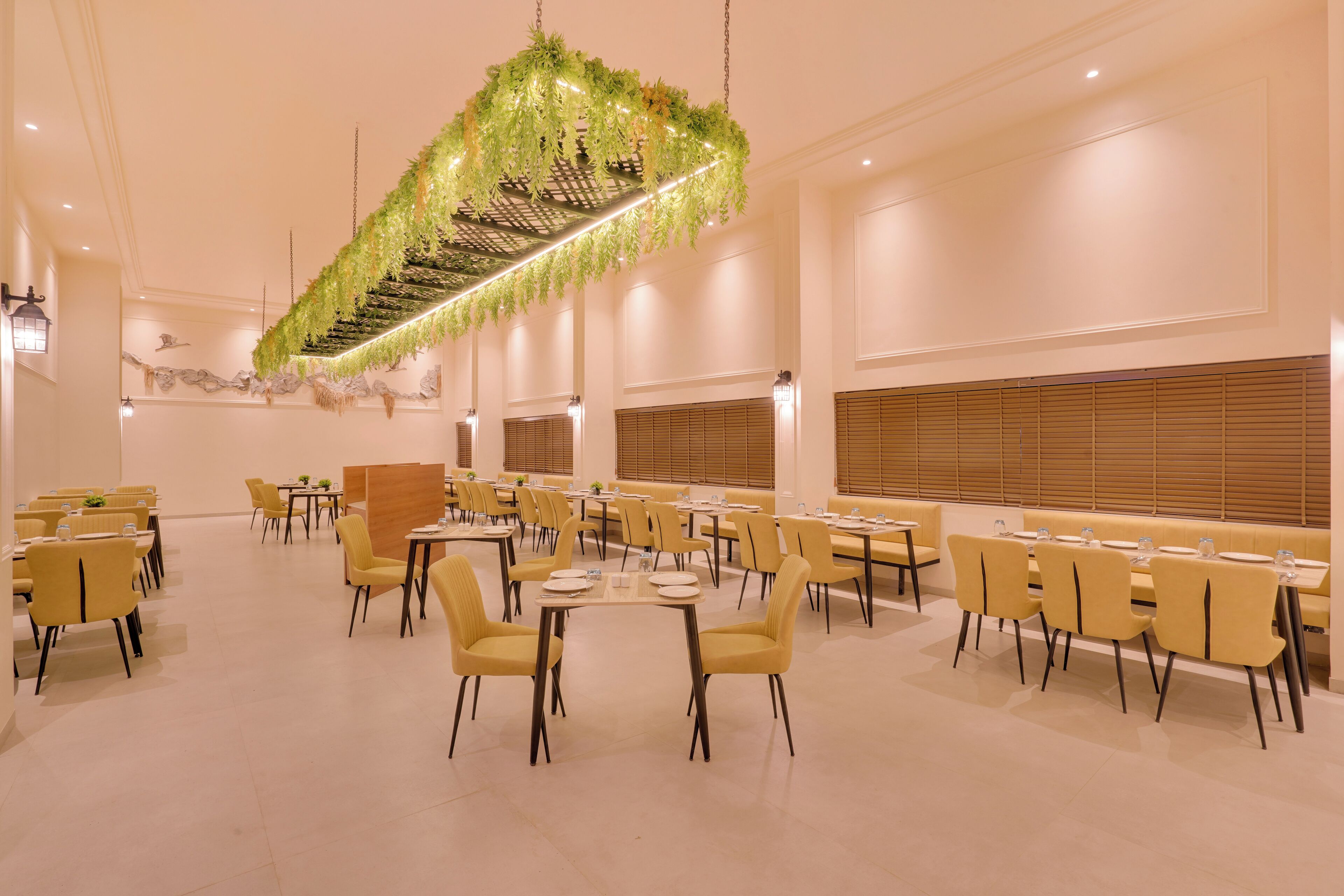 Un vaste espace dédié à la salle à manger, avec plusieurs tables et chaises, une plante décorative suspendue et une rangée de fenêtres équipées de stores.