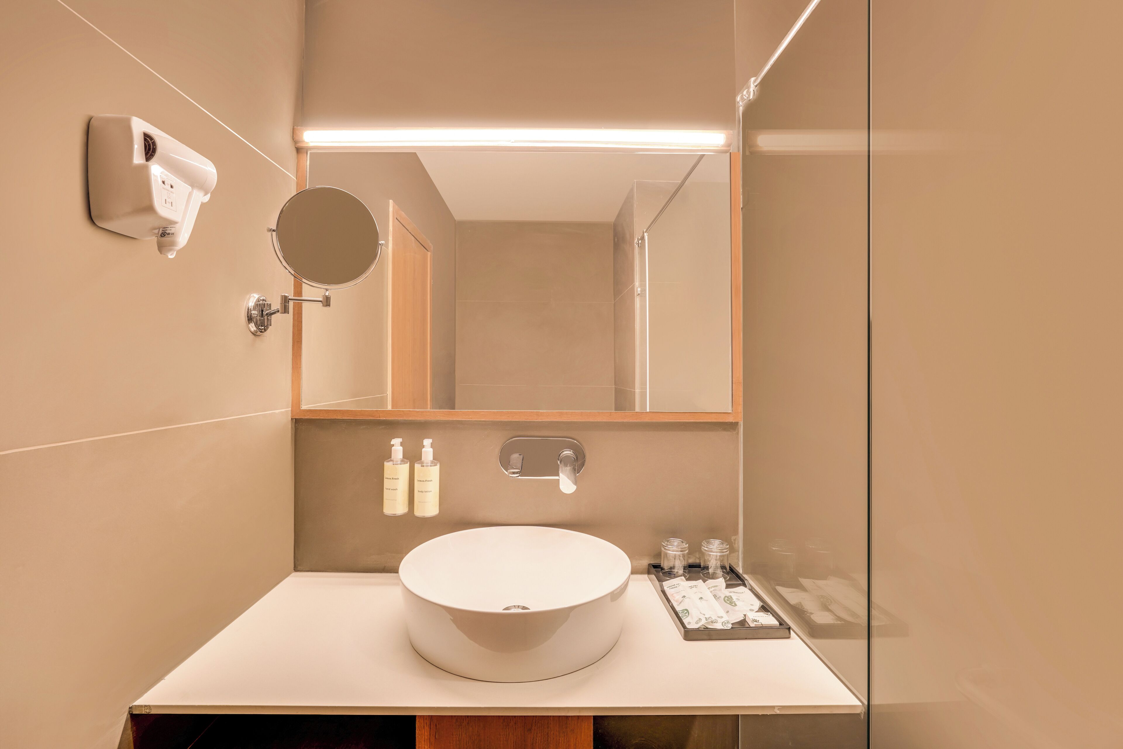 Un baño moderno con un lavamanos redondo, un espejo de mano, dispensador de jabón en la pared y una cabina de ducha con mampara de cristal.