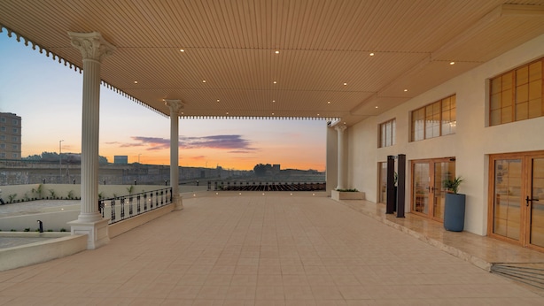 Een ruime overdekte gang met uitzicht op een zonsondergang boven een stadsbeeld, met kolommen en een betegelde vloer.