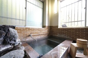 A hot spring bath with a stone wall and tiled floor, a wooden stool, and a window with bars.