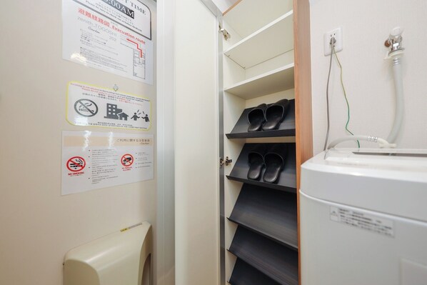 A hotel room with a white wall displaying various safety and emergency information signs, a shoe rack with black shoes, and a white appliance, possibly a refrigerator or heater, on the right side.