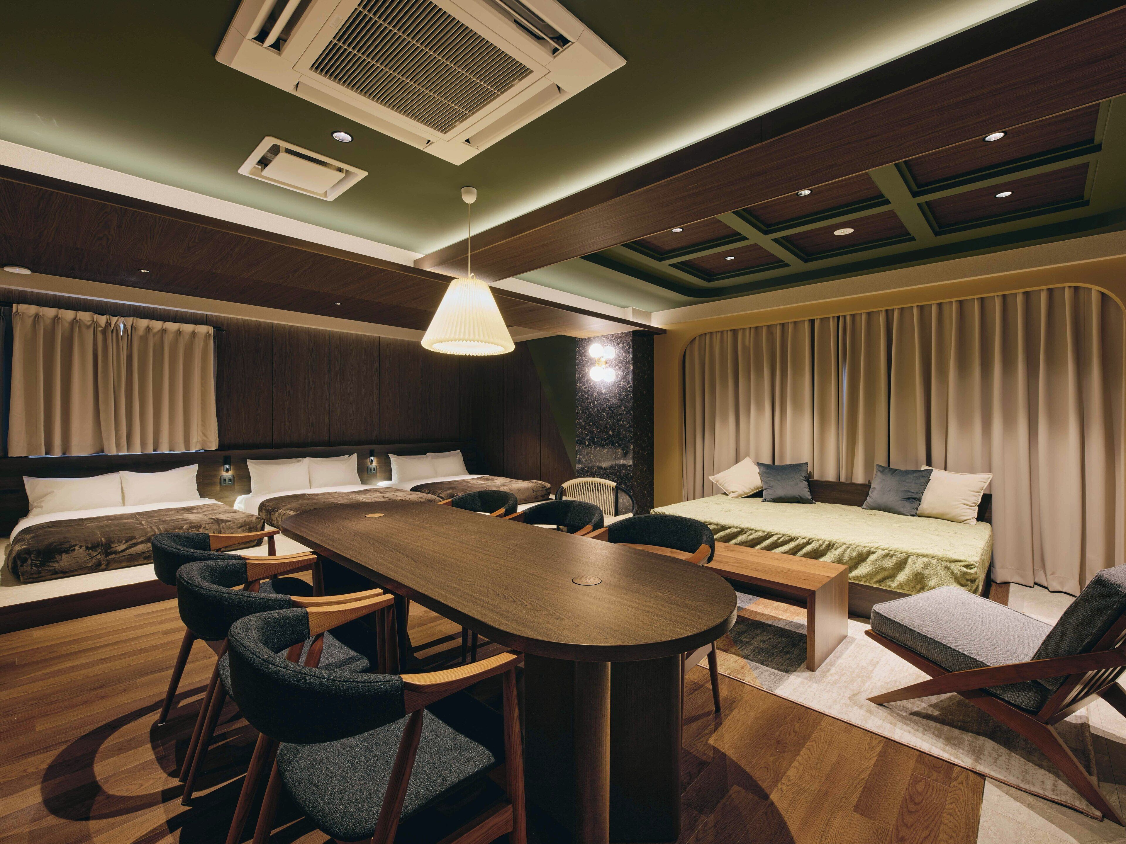 A modern hotel room with a large bed, a dining table, and several chairs. There is a built-in desk and a large window with curtains.