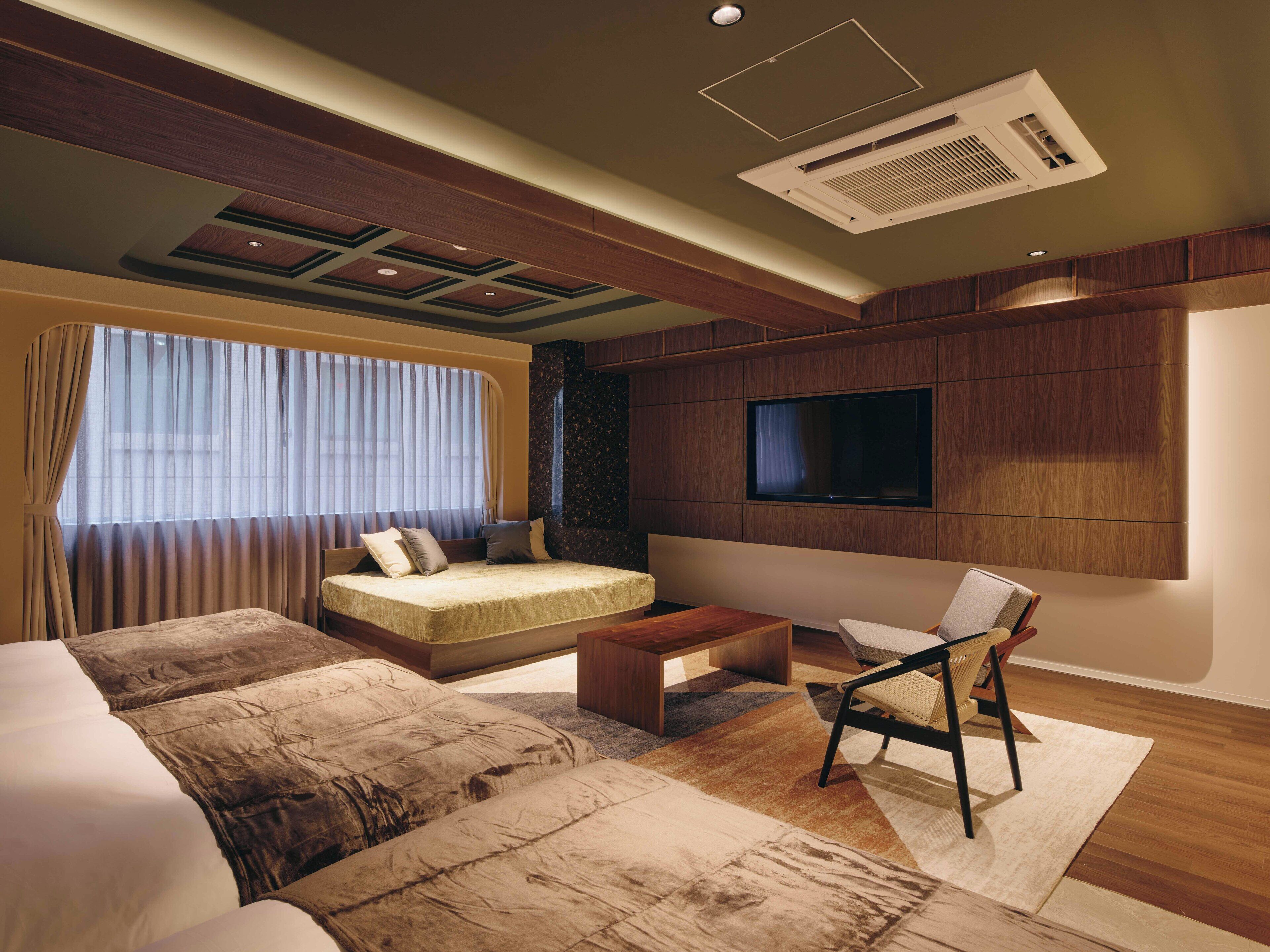 A modern hotel room with a large bed, a sofa, a TV mounted on the wall, and a ceiling with recessed lighting and an air vent.