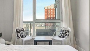 Una habitación de hotel con un ventanal grande, dos sillas blancas con cojines estampados y una mesita.