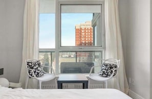Ein Hotelzimmer mit einem großen Fenster, zwei weißen Stühlen mit gemusterten Kissen und einem kleinen Tisch.