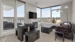 Una sala moderna con televisor grande, muebles oscuros y ventanas amplias que dan a una vista de la ciudad.