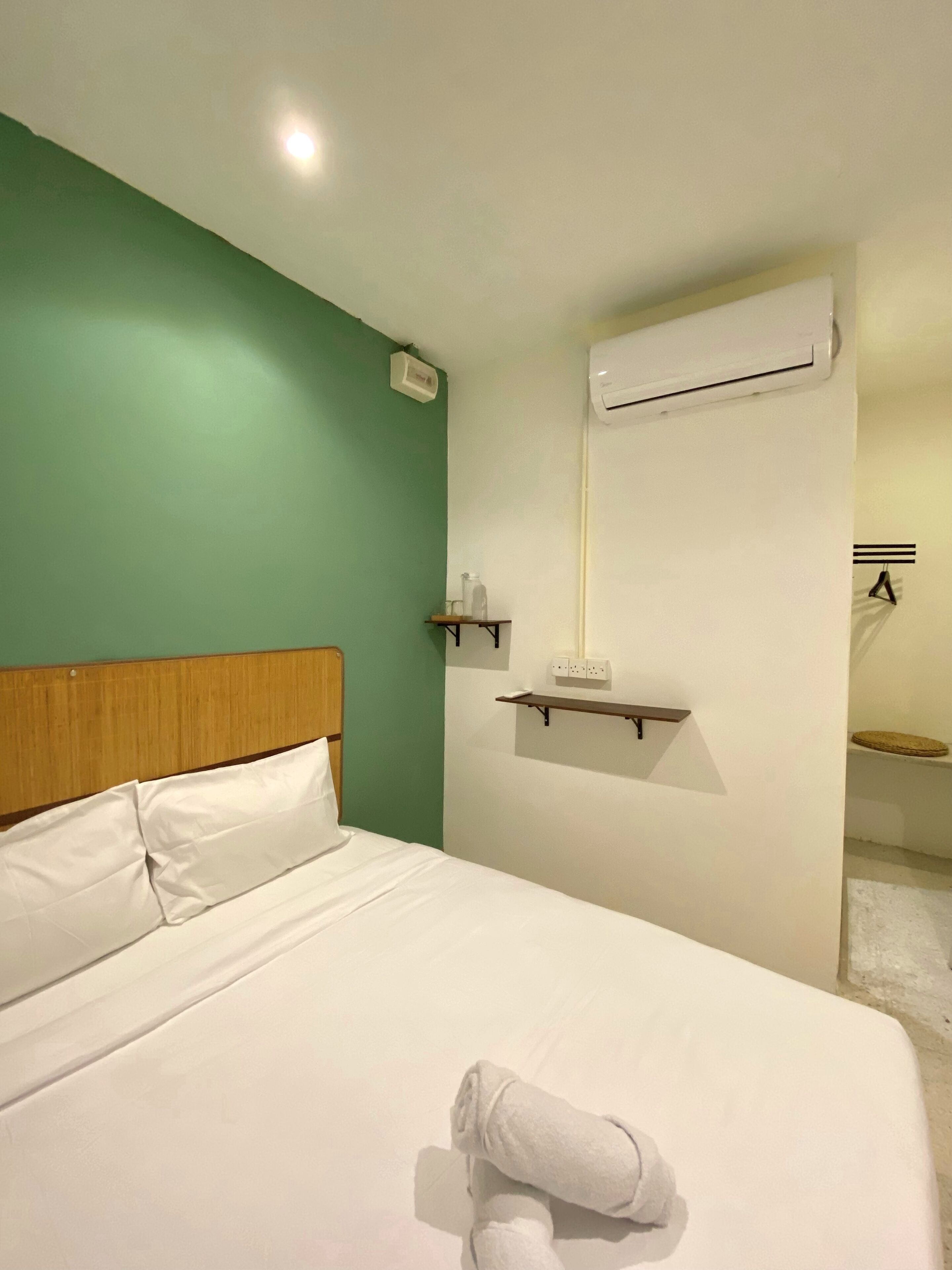 Habitación de hotel con cama, cabecera de madera, pared verde, unidad de aire acondicionado blanca y un pequeño estante con una botella.