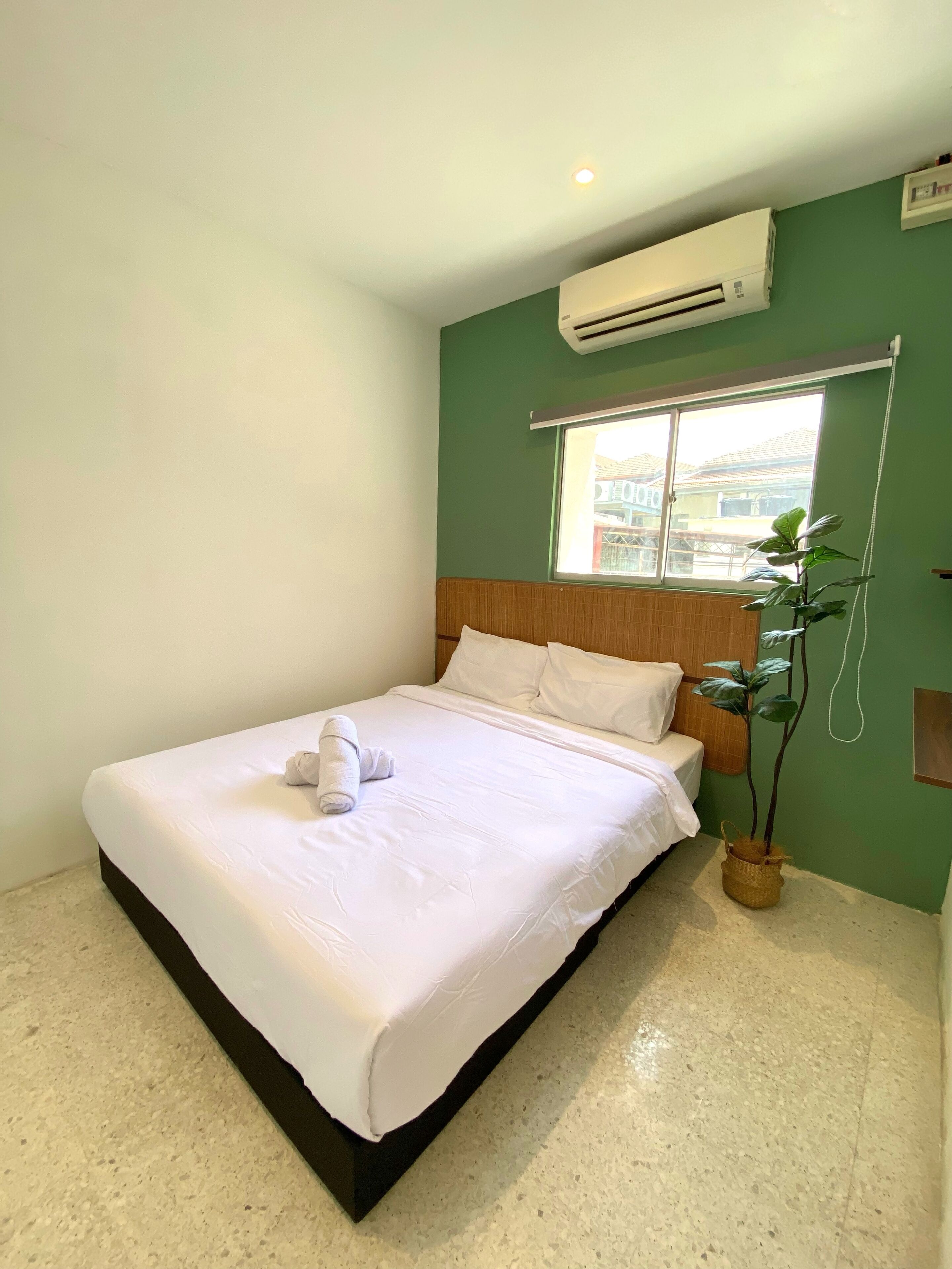 A neatly made bed with white linens and a rolled towel, a potted plant, and a window with a view of buildings outside.