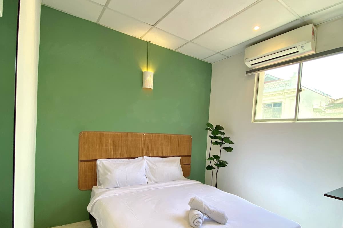 A neatly made bed with white linens and a rolled towel, a wooden headboard, a green wall, a small wall-mounted light, and an air conditioning unit.